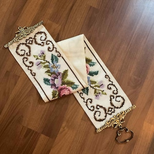 Könnte beinhalten: Cremefarbenes besticktes Textil mit Blumen- und Scroll-Designs. Die Stickerei zeigt rosa, lila, blaue und grüne Blumen. Das Textil hat an jedem Ende verzierte goldfarbene Metallbeschläge mit einer dekorativen Schlaufe.