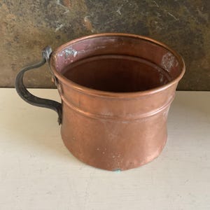 May include: A round copper pot with a wide opening and a single dark metal handle attached to the side. The exterior surface shows a warm metallic finish with subtle signs of wear and oxidation, featuring a smooth, cylindrical body shape.