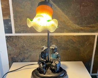 Könnte beinhalten: Dekorative Tischlampe mit einem skulptierten Sockel mit zwei Figuren. Die Lampe hat einen Metallstiel, der einen gerüschten, durchscheinenden Lampenschirm mit Orange- und Grüntönen trägt. Der Sockel ist dunkel, und die Lampe steht auf einer weißen Oberfläche.