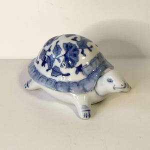 Può includere: Statuina a forma di tartaruga in ceramica con corpo bianco e guscio con motivo floreale blu e bianco. La tartaruga ha una faccia sorridente. Un oggetto decorativo.
