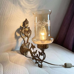 May include: An antique wall sconce with a decorative brass frame and a frosted glass shade. The shade features etched floral patterns and houses a lit bulb. The sconce is mounted on a wall and has a power cord with a plug.