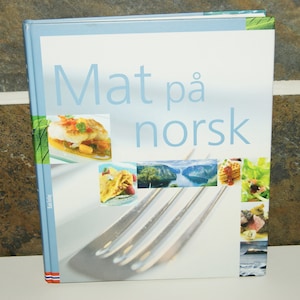 Puede incluir: Un libro de cocina azul y blanco con el título "Mat på norsk" e imágenes de comida y paisajes.