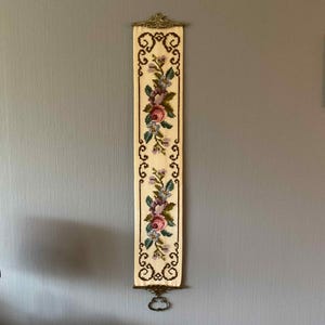 May include: Cream-coloured, embroidered wall hanging with a floral design. The tapestry features pink, purple, and blue flowers with green leaves. It has a decorative metal top and bottom with a hanging loop. The wall hanging is approximately 76 cm long.
