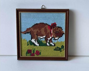 May include: Framed needlepoint art featuring a brown dog with a red bow, set against a blue sky and green grass. The dog has white accents on its paws and is surrounded by red flowers. The frame is made of dark wood.