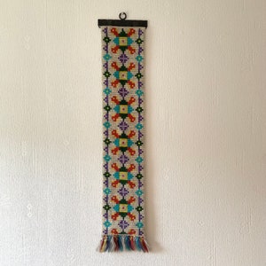 Puede incluir: Un tapiz de pared colorido con un patrón geométrico en tonos de morado, naranja, verde, amarillo y azul. El tapiz está hecho de hilos tejidos y tiene una franja en la parte inferior.