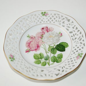 May include: White porcelain plate with a delicate lace design around the edge. The center features a floral design with pink and white roses and green leaves.
