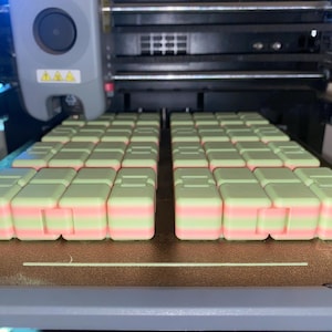 May include: A 3D printer is shown with multiple small, rectangular objects in the process of being printed. The objects are light green with a pink stripe. The printer is gray and black.