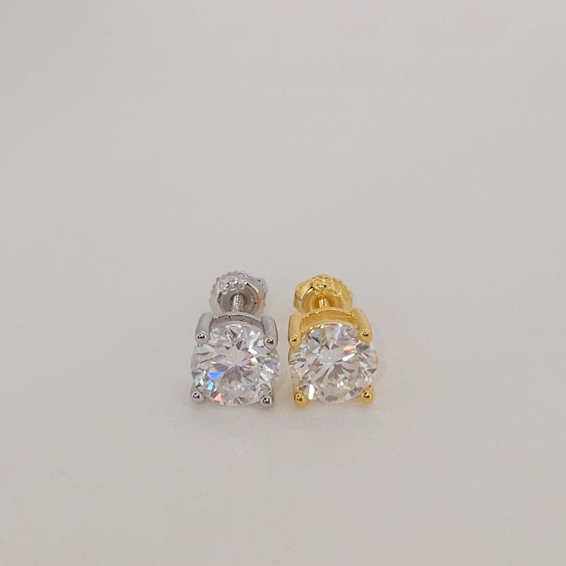 May include: Two stud earrings, one silver and one gold, each with a large, round, clear gemstone. The earrings are displayed against a plain, light background, highlighting their sparkling facets and classic design.