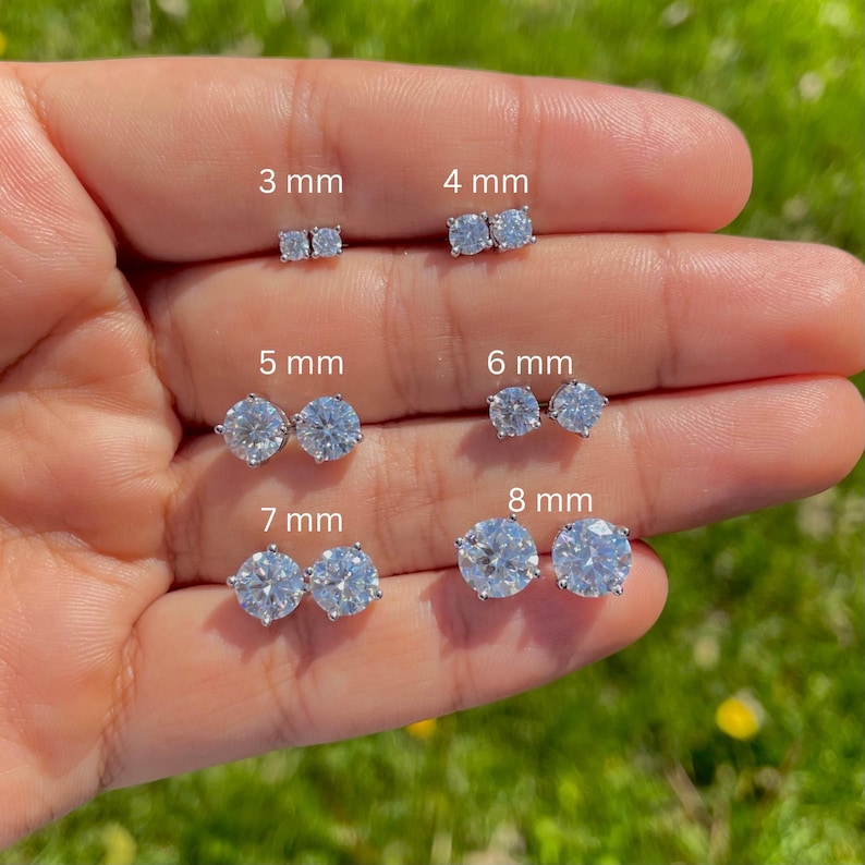 May include: A selection of silver stud earrings in different sizes, from 3 mm to 8 mm, are shown. The earrings are displayed on a hand, with each pair labelled with its size. The clear, sparkling earrings are set against a green backdrop.