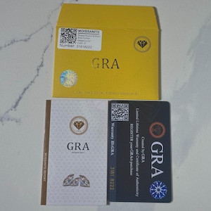 May include: A yellow envelope and two cards with the text "GRA" and "MOISSANITE" are displayed. The envelope has a holographic sticker and a label with details like "Shape Round Brilliant" and "Size 3.0mm". The cards include warranty information.