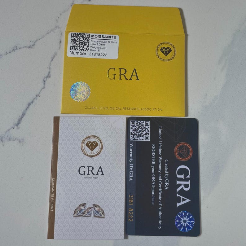 May include: A yellow envelope and two cards with the text "GRA" and "MOISSANITE" are displayed. The envelope has a holographic sticker and a label with details like "Shape Round Brilliant" and "Size 3.0mm". The cards include warranty information.