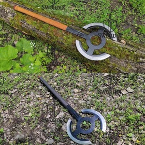 May include: Two axes are displayed. One axe has a wooden handle with a brown leather grip and a black blade with a circular design and silver edges. The other axe has a black handle and blade with a similar design. The axes are on a log and grass.