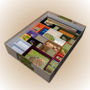 May include: A board game organiser with several compartments in various colours, including orange, purple, and green. The organiser contains game pieces, cards, and a game board. The box is beige with a decorative pattern.