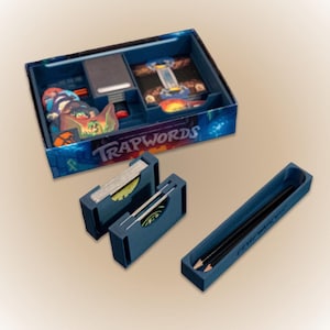 以下が含まれることがあります： 青い箱に入ったボードゲーム「TRAPWORDS」。箱にはゲームカード、トークン、小さな本が入っています。カードホルダー2つと鉛筆トレイも確認できます。