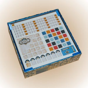 Può includere: Una scatola di gioco da tavolo con la parola "AZUL" stampata sul tabellone. Il tabellone presenta una griglia, tessere colorate e aree di punteggio. La scatola ha un bordo blu e un esterno a motivi.