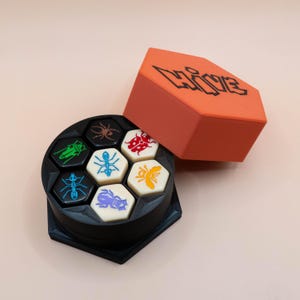Puede incluir: Un tablero de juego circular negro con espacios hexagonales, cada uno con una ilustración de insecto diferente. Las piezas del juego son hexágonos blancos con diseños de insectos. Una caja hexagonal naranja con la palabra "HIVE" en letras de grafiti negras está abierta.