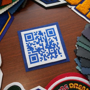 May include: A close-up of a white patch with a blue embroidered QR code surrounded by a blue border. The patch is on a wooden surface with other patches in various colors and designs.