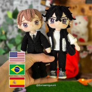 May include: Two crocheted dolls with large eyes and detailed faces. One doll is in a black suit, the other in a black and white outfit. The dolls are held in a hand, with flags of the USA, Brazil, and Spain visible.