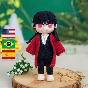 May include: Crocheted amigurumi doll with black hair, red and black clothing, and red eyes. The doll is standing on a wooden surface with a gold chain in the background.
