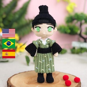 Op de afbeelding: Een gehaakte pop die een groene en zwarte kimono draagt met een zwarte haarknot. De pop heeft groene ogen en een lichte huidskleur. De pop staat op een houten oppervlak met rode pompoenen.