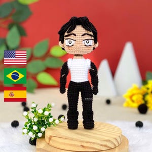 Puede incluir: Muñeco de ganchillo hecho a mano de un personaje de anime masculino. El muñeco tiene pelo negro, cara morena y viste una camisa blanca con mangas negras y pantalones negros. El muñeco está sobre una pequeña plataforma de madera. El fondo es rojo.