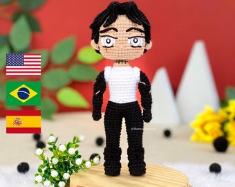 May include: A handmade crochet doll of a male anime character. The doll has black hair, a tan face, and is wearing a white shirt with black sleeves and black pants. The doll is standing on a small wooden platform. The background is red.