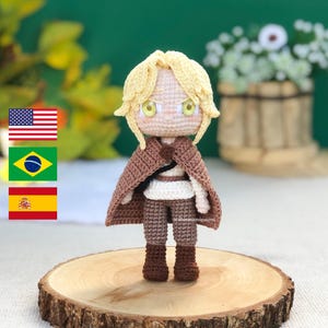 Pode incluir: Boneca de crochê feita à mão com penteado loiro, usando capa e calças marrons e botas marrons. A boneca tem uma camisa branca e está em pé em uma fatia de madeira. A boneca tem olhos amarelos.