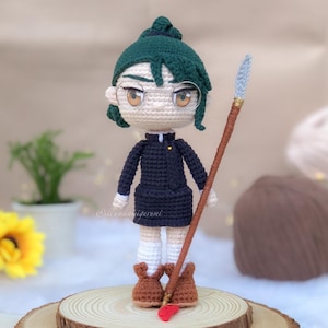 Puede incluir: Muñeca amigurumi de crochet con cabello verde, vistiendo un vestido negro y zapatos marrones, sosteniendo un palo de madera con una punta azul.