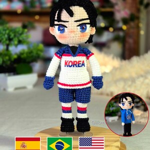 Peut inclure: Une poupée au crochet avec des cheveux noirs et des yeux bleus, portant un maillot blanc avec le mot KOREA en bleu et rouge, un short bleu et des chaussures noires. Un petit encart circulaire montre la même poupée avec un sweat bleu.