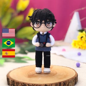 Puede incluir: Una muñeca amigurumi de crochet de un niño que lleva un chaleco de punto azul, pantalones negros y zapatos blancos. Está sosteniendo una cámara y lleva gafas. La muñeca está de pie sobre una superficie de madera.