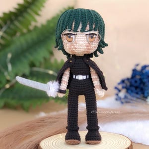 Pode incluir: Figura amigurumi de crochê de um personagem com cabelo verde, vestindo roupas pretas e óculos, segurando uma espada prateada. A figura está em pé sobre uma superfície de madeira.