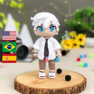 Pode incluir: Uma boneca de crochê com cabelo branco, uma camisa branca, calças marrons e sapatos brancos. A boneca está em pé sobre uma superfície de madeira com objetos redondos pretos.