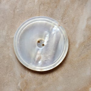 May include: A large, round, pearlescent button with two small holes. The button has a subtle iridescent sheen and a raised outer rim. The button is likely made of mother-of-pearl and is suitable for sewing on garments or crafts.