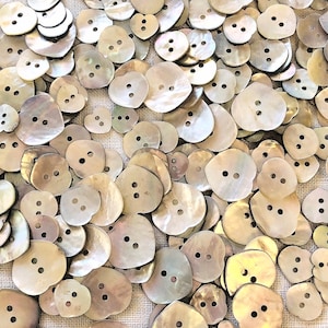 May include: A collection of iridescent, mother-of-pearl buttons in various sizes and shapes. The buttons are a pale, pearlescent white with a subtle green hue. Some buttons are heart-shaped, while others are round or oval.