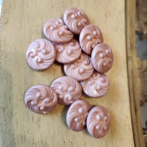 May include: A collection of eleven round, dusty pink buttons. Each button features a raised, swirling relief pattern that curves across the surface, accented by small, smooth circular dots integrated into the textured, fan-like design.