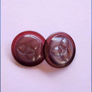 May include: Two round, dark burgundy buttons with a raised, three-petal floral design. The buttons are made of a glossy material and are set against a light pink background.