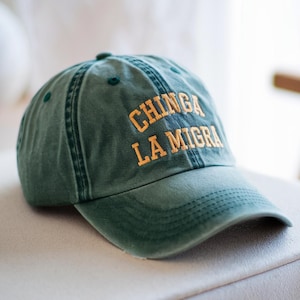 May include: A green baseball cap with the text "CHINGA LA MIGRA" embroidered in yellow. The cap has a curved brim and a distressed, vintage look. The hat is sitting on a white surface.