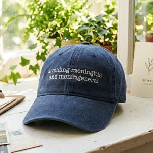 May include: A navy blue baseball cap with the words "avoiding meningitis and meningeneneral" embroidered in white. The cap is sitting on a white surface near a window with plants and a postcard.