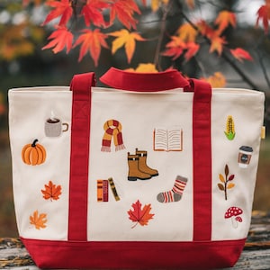 May include: Cream-colored tote bag with red accents and handles, adorned with embroidered fall-themed patches. Patches include a pumpkin, coffee cup, scarf, boots, books, leaves, and a mushroom. The bag is set against a backdrop of autumn foliage.