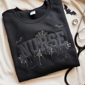May include: Black sweatshirt with the word "NURSE" in gray, featuring a spiderweb and dripping design. Bats and a stethoscope are also visible. The sweatshirt is folded on a cream-colored surface.