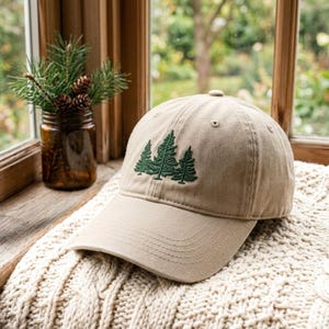 May include: A beige baseball cap featuring an embroidered design of three dark green pine trees on the front panel. The cap has a curved brim and stitched eyelets, showcasing a structured crown construction with a classic six-panel design.
