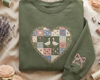 Peut inclure: Un sweat-shirt vert olive avec un motif patchwork en forme de cœur représentant deux oies. Le cœur est bordé d'un bord festonné. La manche droite porte un nœud rose et le nom "Alice" brodé. Des fournitures de couture et des fleurs entourent le sweat-shirt.