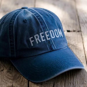 Puede incluir: Gorra de b&eacute;isbol azul marino con la palabra "FREEDOM" bordada en blanco. La gorra est&aacute; hecha de un material suave y tiene una visera curva. El sombrero est&aacute; sobre una superficie de madera.