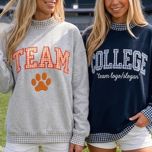 May include: A light gray sweatshirt with the word TEAM in orange gingham letters and an orange paw print, and a navy blue sweatshirt with the word COLLEGE in blue gingham letters and the text team logo/slogan, both with gingham trim.