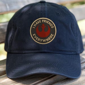 May include: Navy blue baseball cap with a circular emblem featuring the text "I HAVE FRIENDS EVERYWHERE" around a red symbol. The cap is made of a soft material and has a curved brim.