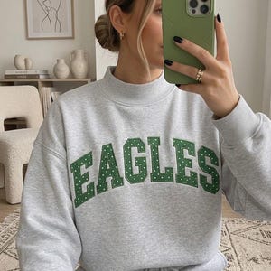 May include: A light gray crewneck sweatshirt featuring a mock neck collar and long sleeves. The front displays the word EAGLES in large, dark green, block-style letters with a white polka dot pattern, stitched in an arched formation across the chest.