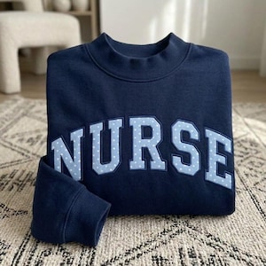 May include: A dark navy blue crewneck sweatshirt featuring the word NURSE in large, arched, light blue letters. Each letter has a white polka dot pattern and a dark navy blue outline, creating a bold, layered effect on the front of the garment.