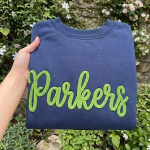 May include: A navy blue crewneck sweatshirt featuring the word Parkers embroidered in a bright lime green cursive font across the chest. The garment has a ribbed neckline and long sleeves, showcasing a clean design with bold, textured lettering.