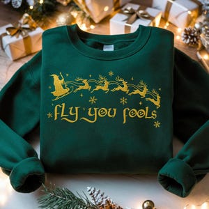 May include: A dark green sweatshirt with gold text and graphics. The design features a witch on a broomstick being pulled by reindeer, snowflakes, and the phrase "fly you fools". The sweatshirt is a festive holiday item.
