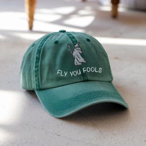 May include: A green baseball cap with the embroidered phrase "FLY YOU FOOLS" and a wizard graphic. The cap is made of a soft material and has a curved brim. The hat is a solid green color.